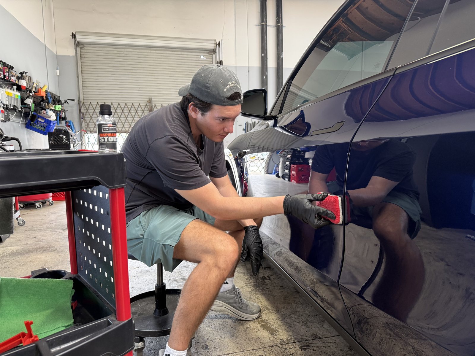 Scott on a rolling stool working an applicator along the lower door of a dark blue SUV, his reflection showing in the paint